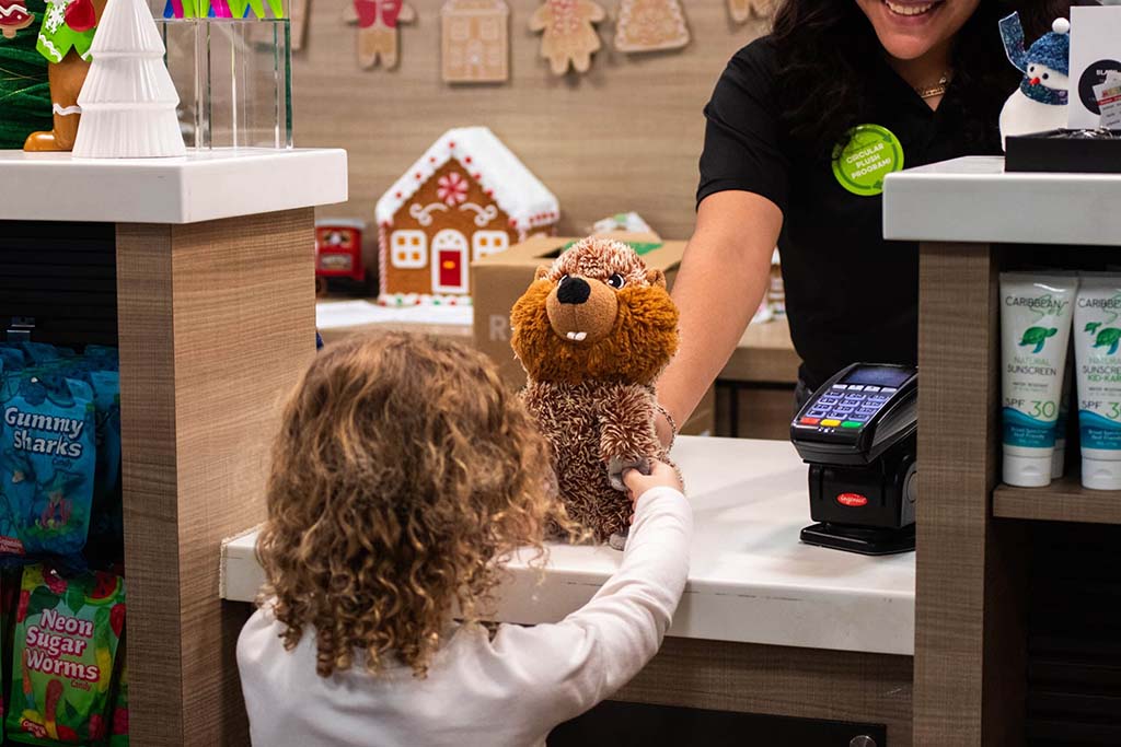 A child with curly hair hands a brown plush toy to a smiling cashier at a store counter decorated with holiday items and candy displays.
