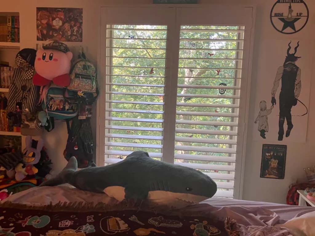 A bedroom with a large shark plush toy on the bed, surrounded by anime posters, plush toys, and decorative shelves near a sunlit window.