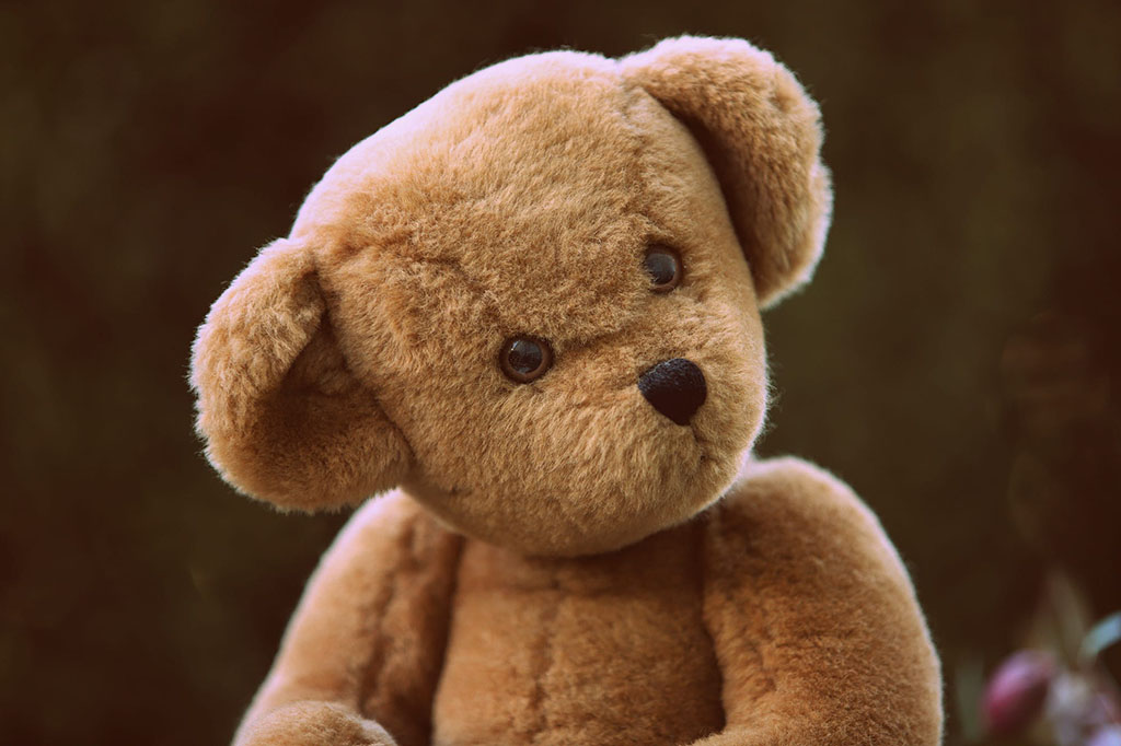 Soft brown teddy bear with black button eyes and nose, slightly tilting its head against a dark blurred background, showcasing a traditional plush toy design.