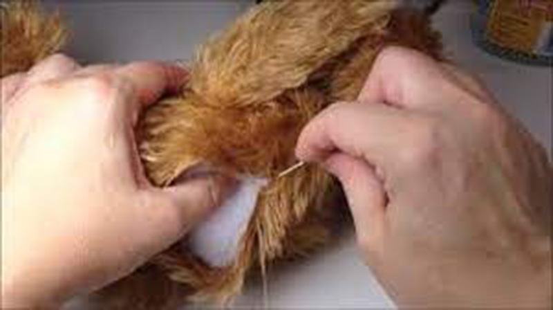 Hands repairing a plush toy by hand sewing an opening closed around the stuffing.