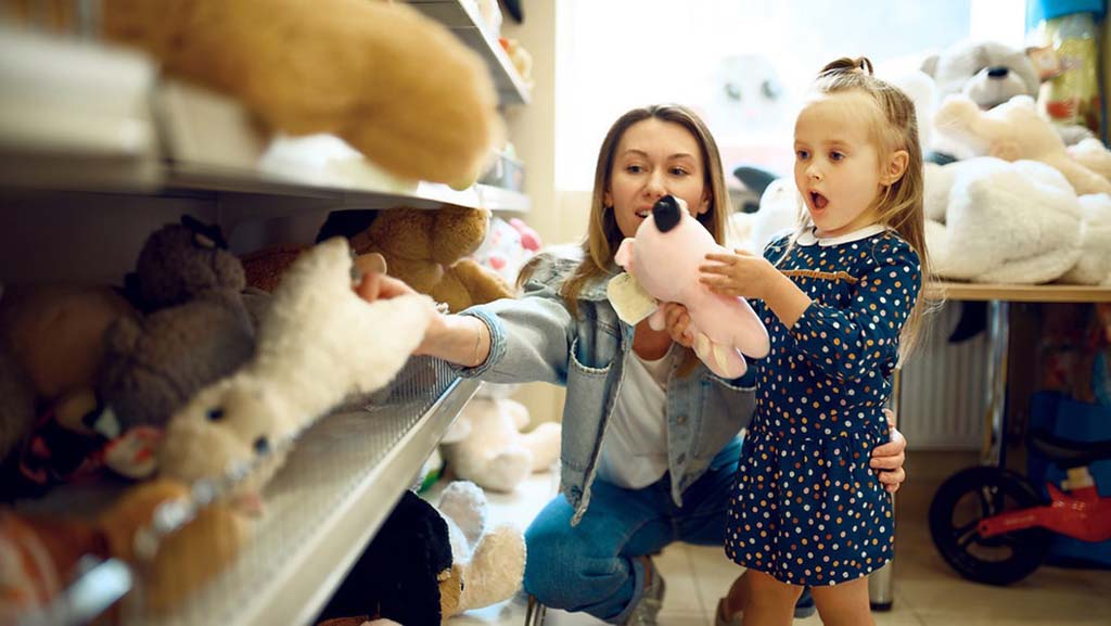 A mother and young girl choosing stuffed animals together in a toy store, with the child excitedly holding a pink plush toy.