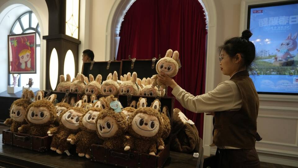 A store employee arranges a large display of brown, fluffy plush toys with big smiling faces and bunny-like ears, all stacked in open vintage-style trunks inside a well-lit retail shop.