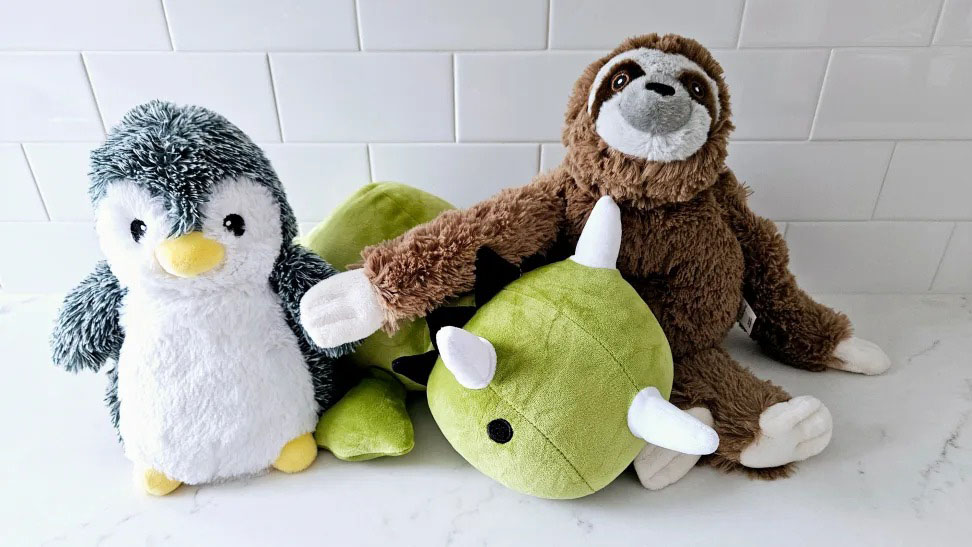A cute group of plush toys including a fluffy penguin, a brown sloth, and a green dinosaur with white horns, displayed together on a white countertop in front of a tiled wall.