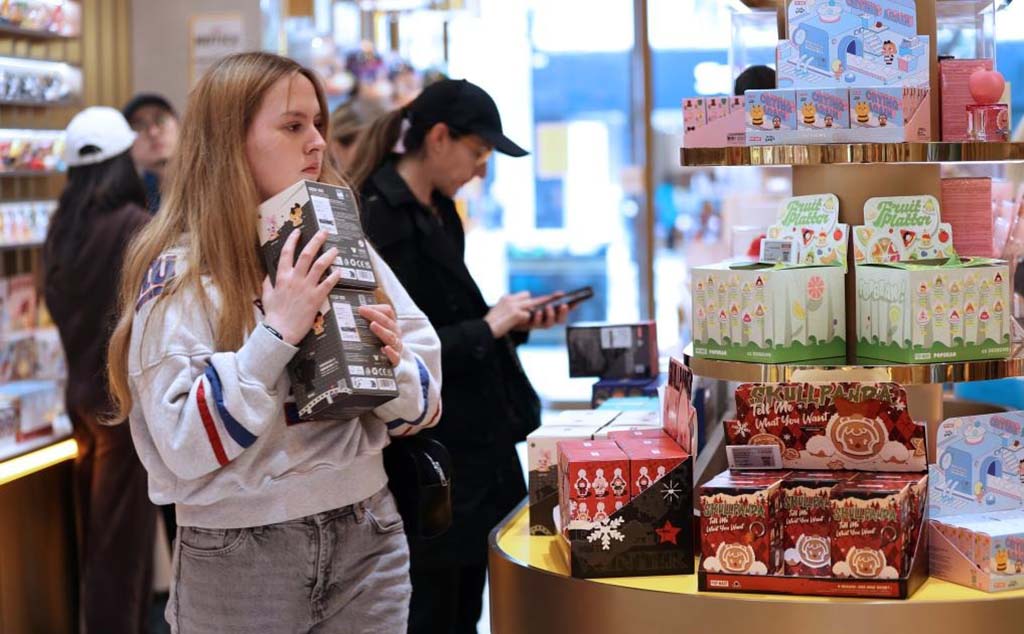  People browsing collectible blind box toys in a brightly lit designer toy store, with one woman holding several boxes near display shelves.