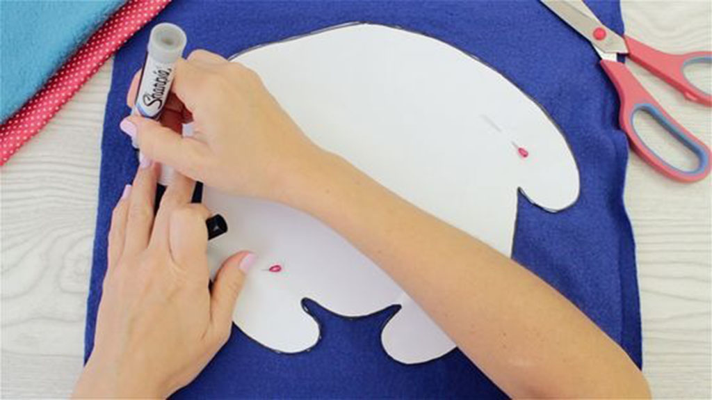 Hands tracing a plush toy sewing pattern onto blue fabric using a marker, with scissors and pins nearby for cutting and assembly.