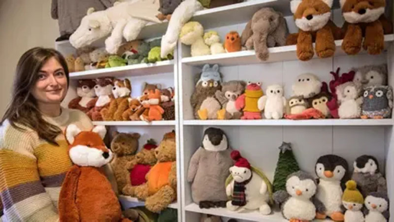 A woman standing beside shelves filled with a large variety of plush animals, including foxes, owls, penguins, elephants, and teddy bears, all organized neatly in a display.