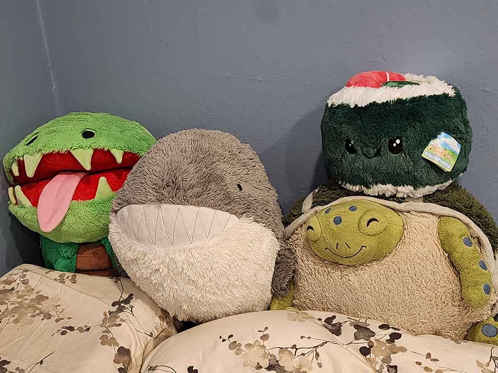 A group of four plush toys—an open-mouth green crocodile, a round gray shark, a sushi roll Squishmallow, and a smiling turtle—sit together on a bed against a blue wall.