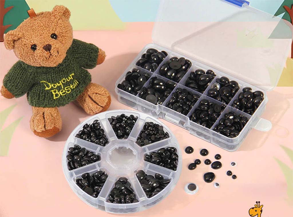 Assorted black plastic safety eyes in organized storage boxes placed beside a small brown teddy bear wearing a green sweater.
