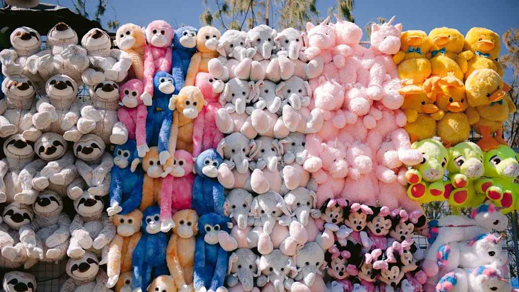 A vibrant wall of stuffed animals including monkeys, elephants, sloths, ducks, and unicorns, neatly arranged in rows at an outdoor fair.