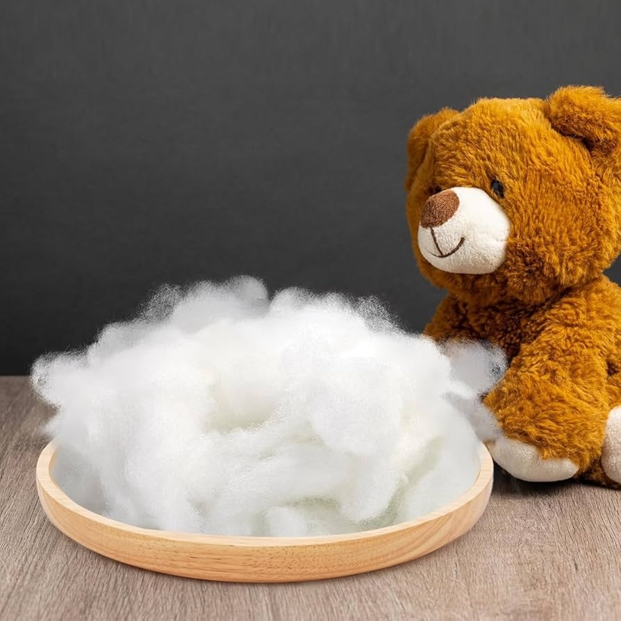 A brown plush teddy bear sits beside a wooden tray filled with soft white polyester fiber stuffing, placed on a wooden surface with a dark background.