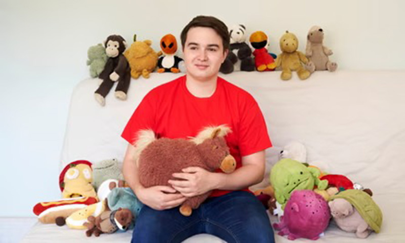 A young man in a red shirt sits on a couch holding a brown plush horse, surrounded by various stuffed animals placed beside him and on the backrest.