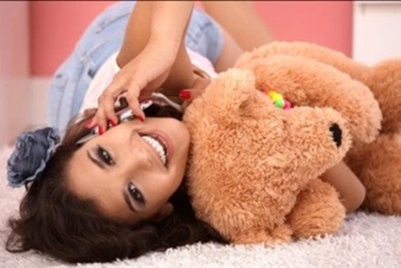 A smiling young woman lies on a soft carpet while talking on the phone and hugging a large brown teddy bear in a cozy pink bedroom.