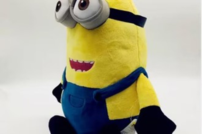 A yellow plush toy with goggles, black gloves, and blue overalls, sitting upright against a plain white background.