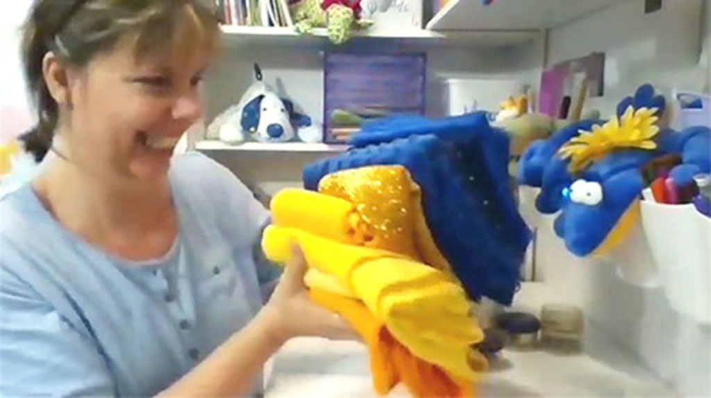 Smiling woman holding colorful yellow and blue fabrics in a sewing room, selecting materials for handmade plush toy projects.
