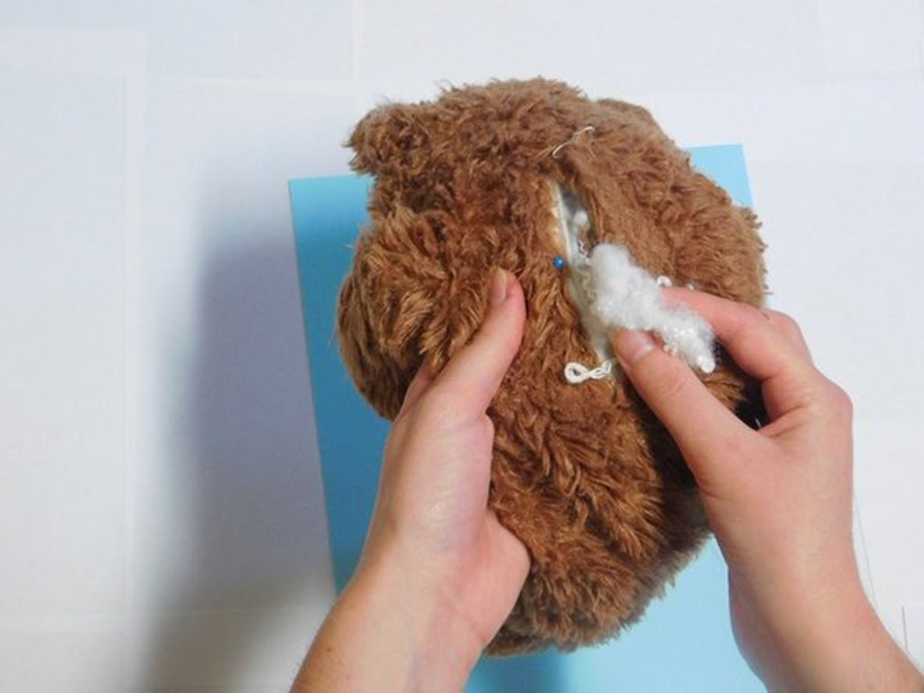 Hands inserting new polyester fiberfill into an open seam of a brown teddy bear during a plush toy repair.