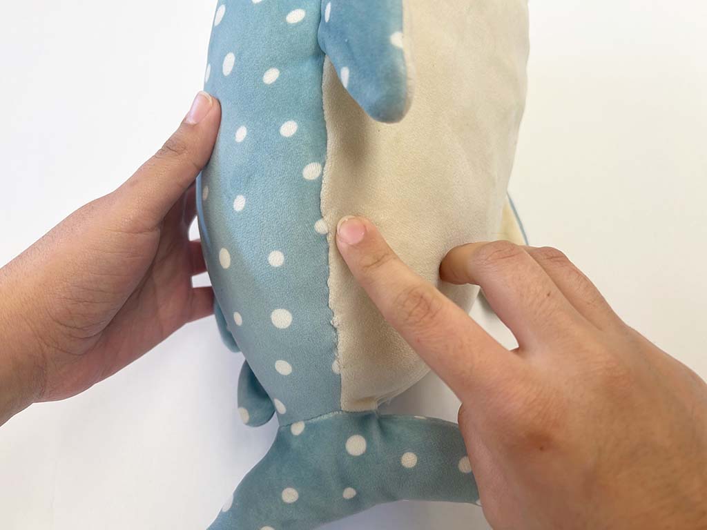 Hands inspecting the side seam opening of a blue polka-dot plush toy, showing where the stuffing area is located before closing the seam.