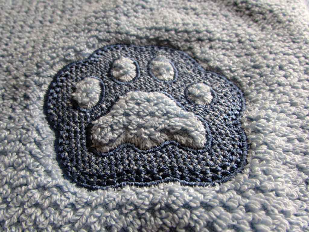 Close-up of a blue embroidered paw print design stitched onto a textured terry fabric, showing raised and flat embroidery details.