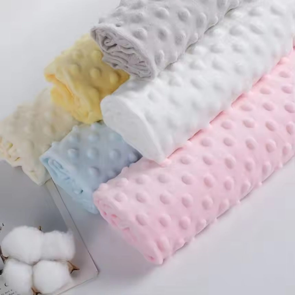 Soft pastel-colored dimple minky fabric rolls in pink, blue, white, yellow, and grey arranged neatly, showcasing their raised dot texture and plush surface.