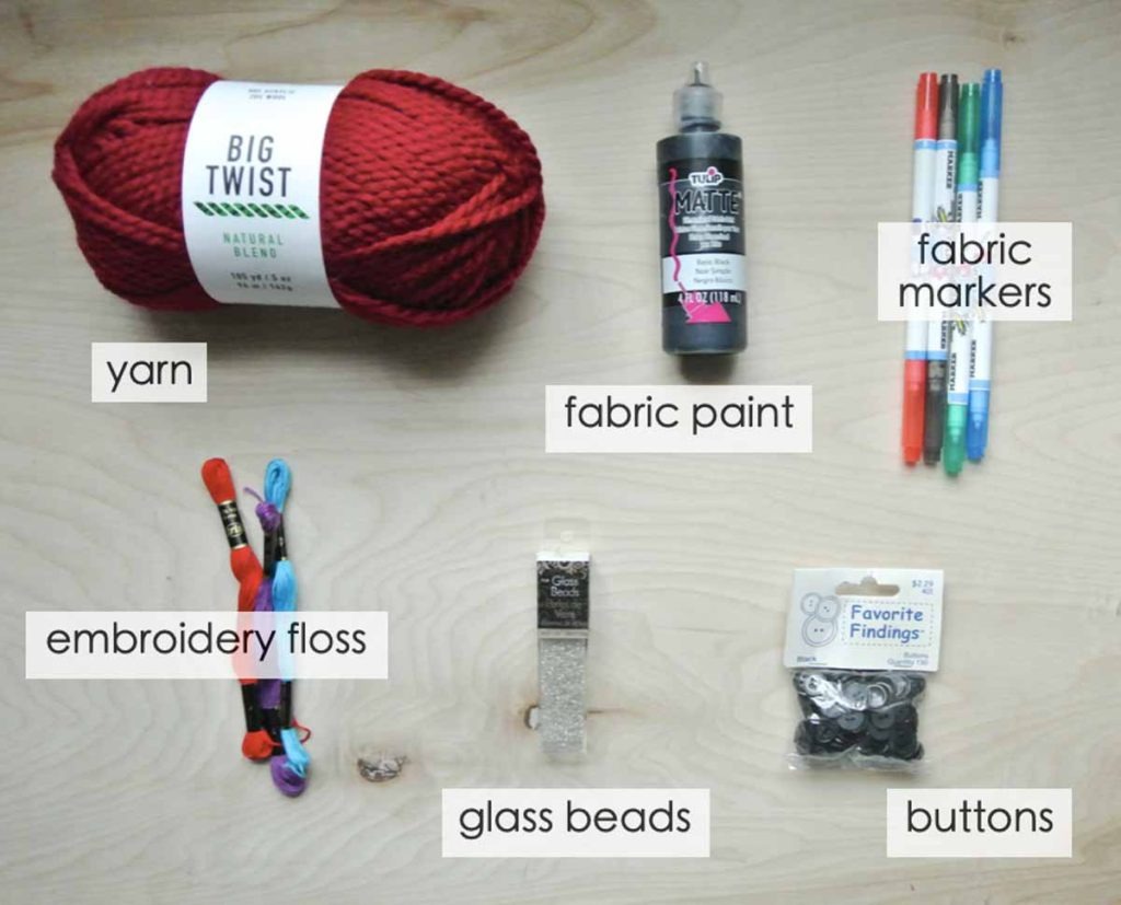Various plush-making materials including yarn, embroidery floss, fabric paint, fabric markers, glass beads, and buttons neatly arranged on a wooden table with labels.