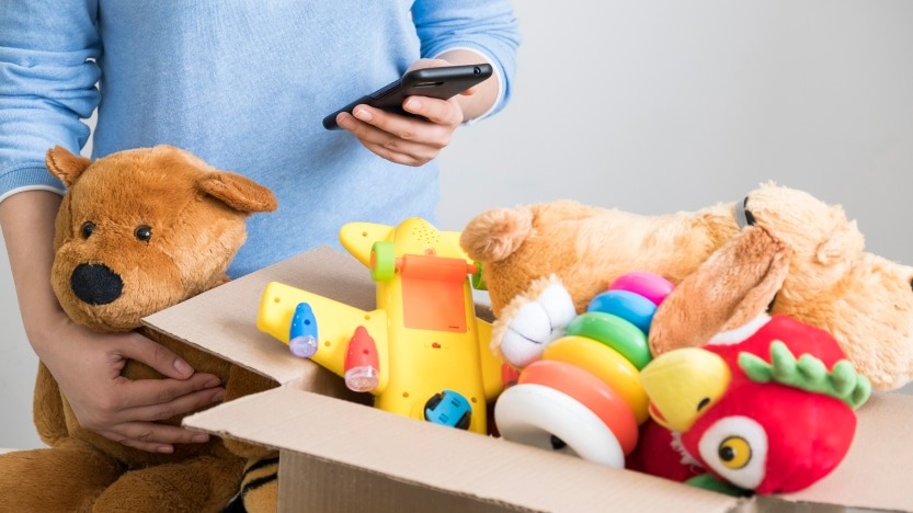 A person holding a phone while packing a cardboard box full of children’s toys, including plush animals, a colorful stacking ring set, and a yellow toy airplane.