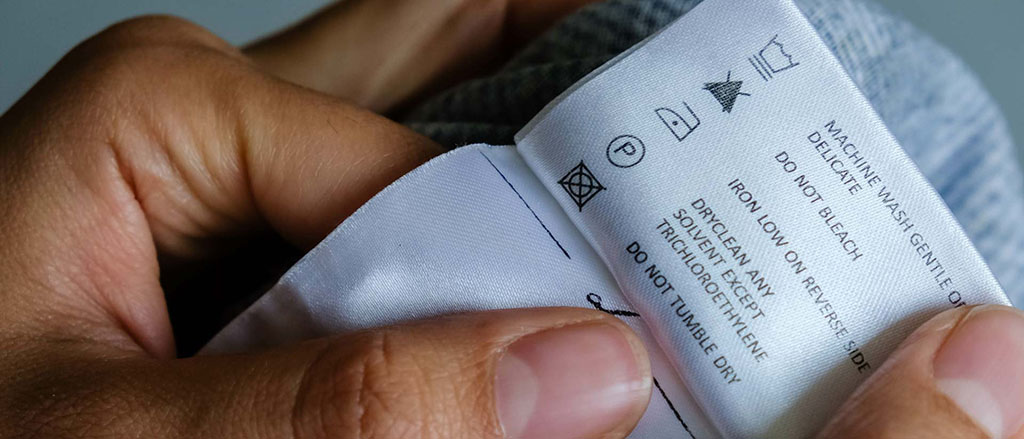 Close-up of hands holding a textile care label showing washing, ironing, and drying instructions, commonly used for clothing, plush toys, and fabric compliance labeling.