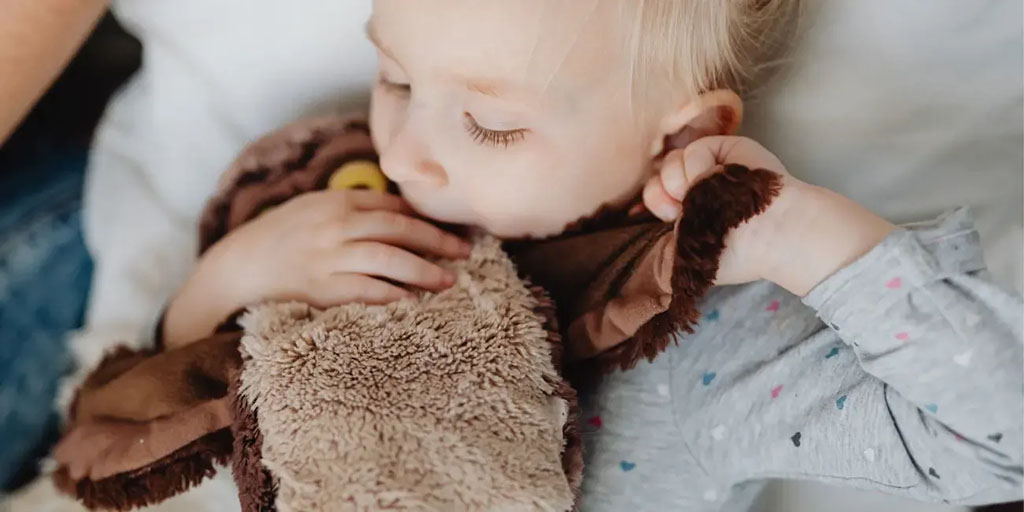 A small child lies in bed, gently holding and snuggling a soft brown owl plush toy close to their face, creating a calm and comforting bedtime moment.