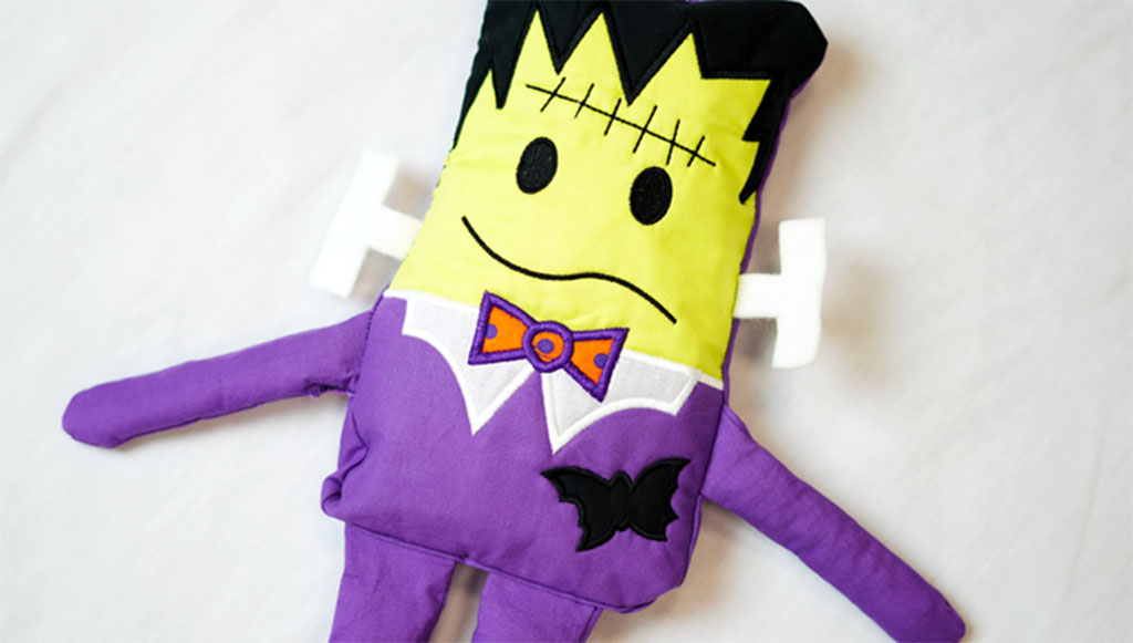 A handmade Frankenstein-themed plush toy with a yellow face, purple outfit, bow tie, and stitched details lying on a white surface.
