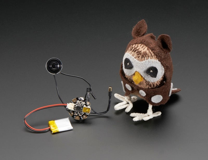 A small brown plush owl placed beside loose electronic voice-module components, including a speaker, circuit board, and battery, showing how sound features can be added to stuffed animals.