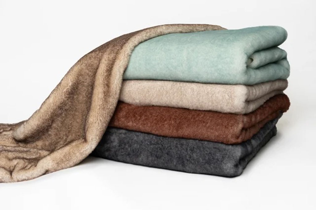 Stacked pieces of soft plush fabric in multiple colors, including beige, brown, grey, and mint green, used for making high-quality stuffed animals and plush toys.