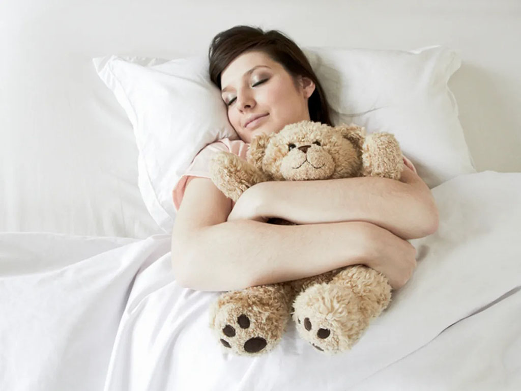 A woman peacefully sleeps in bed hugging a soft brown teddy bear, surrounded by white pillows and bedding.