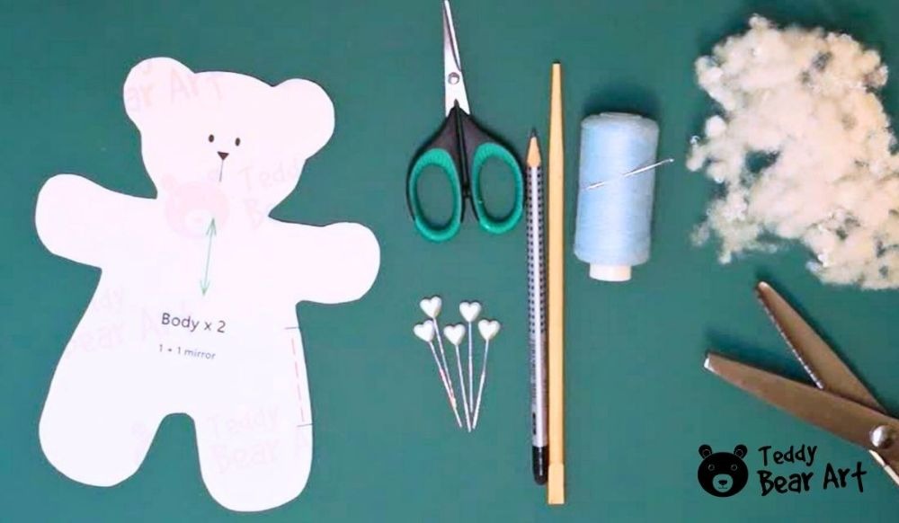 A teddy bear sewing pattern laid out alongside crafting tools including scissors, pins, pencil, thread, and stuffing on a green workspace background.