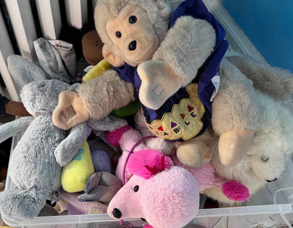 A clear storage bin filled with assorted stuffed animals, including a monkey plush wearing a vest, a pink mouse plush, a grey bunny plush, and several other soft toys piled together.