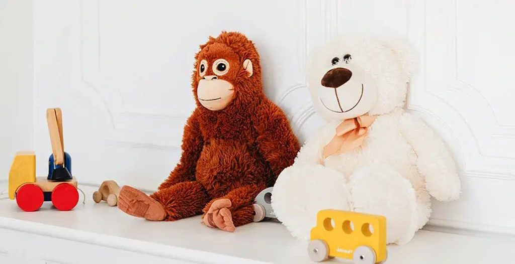A brown monkey plush and a white teddy bear with an orange ribbon sitting on a white shelf beside colorful wooden toy cars.