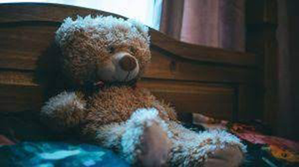A soft brown teddy bear sitting upright on a wooden bed in a dimly lit bedroom.