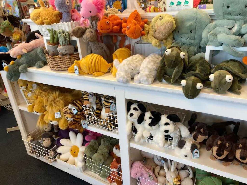 A display of various plush toys arranged on white shelves, including turtles, octopuses, cats, dogs, flowers, and other stuffed animals in a toy shop.