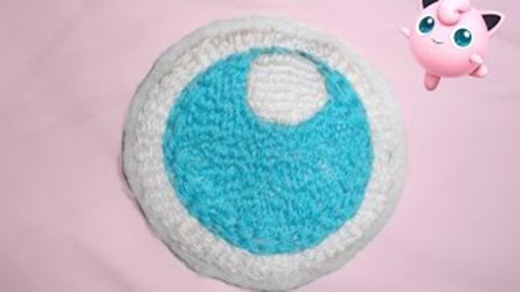 A crocheted round patch designed to look like a large blue and white anime-style eye, placed on a pink background with a small illustration of a pink cartoon character in the corner.
