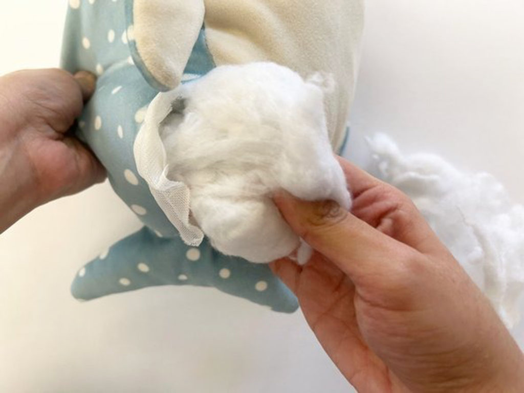  Hands inserting white polyester cotton filling into a blue polka-dot stuffed animal through an open seam during plush repair.