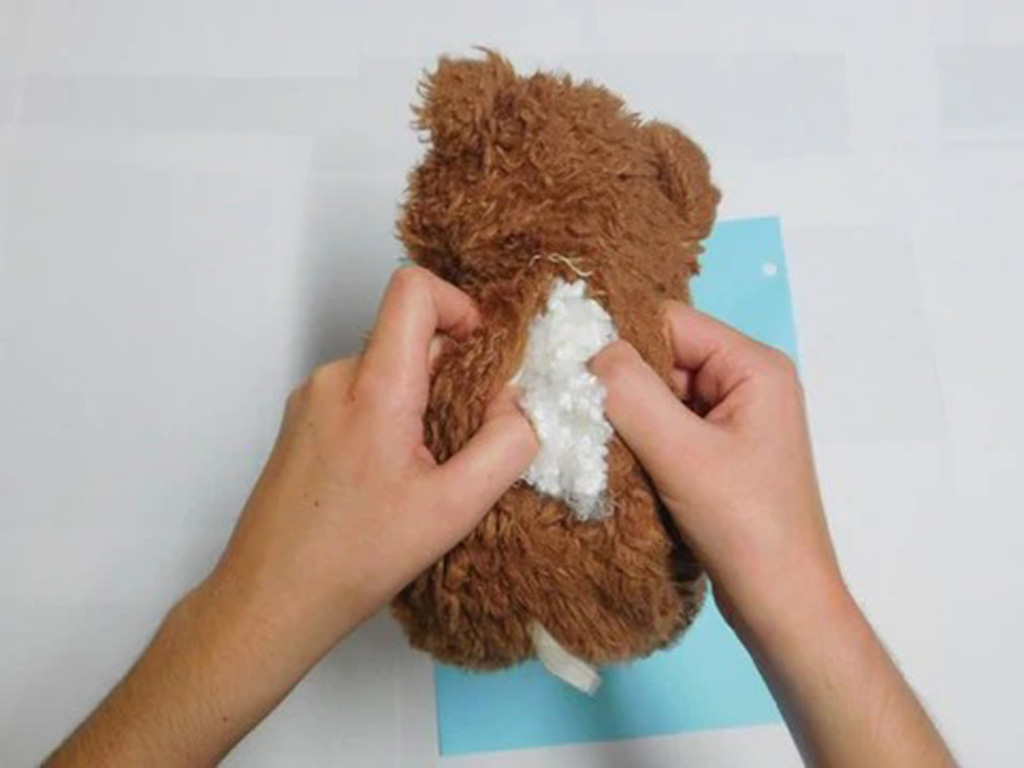 Hands opening the back of a brown stuffed toy to adjust or add white stuffing material inside.