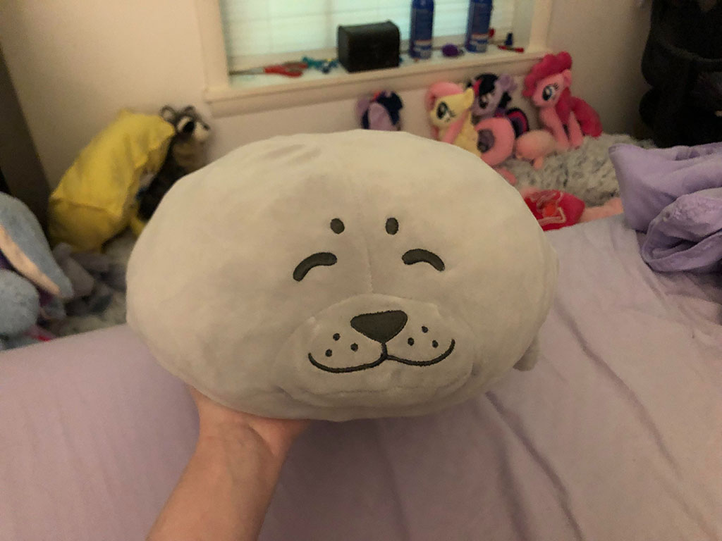 Hand holding a round gray plush seal with a smiling embroidered face, resting on a bed surrounded by other stuffed animals.