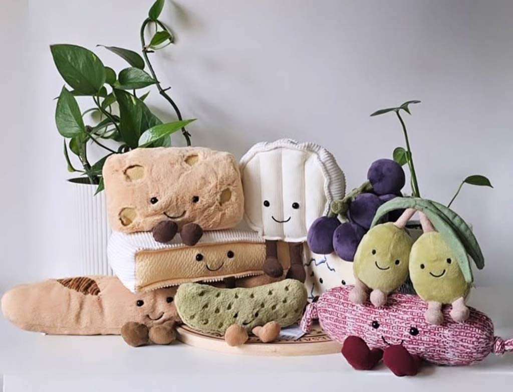 “A group of soft plush food toys arranged on a white surface, including smiling bread slices, grapes, olives, and other character-themed plushies, positioned beside green leafy plants for decoration.”