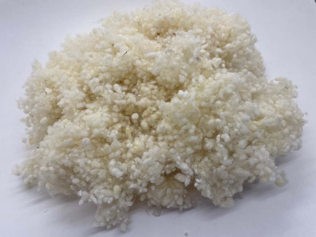 A close-up pile of off-white natural wool stuffing fibers on a plain surface, showing its soft, fluffy, and textured appearance used for plush toy filling.