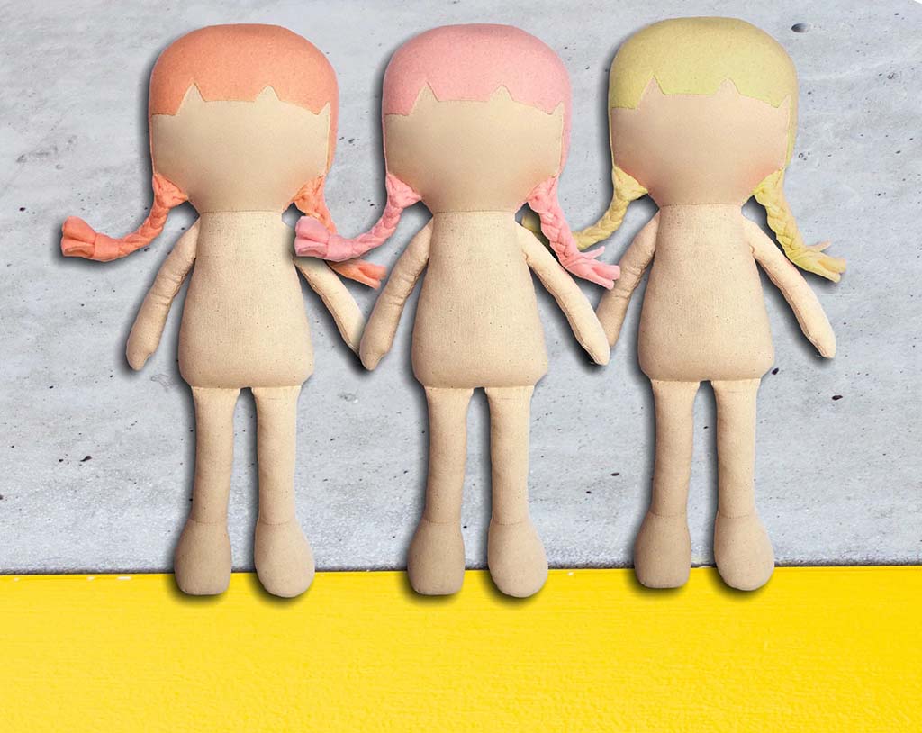 Three blank cloth plush dolls with braided hair—one orange, one pink, and one yellow—stand side by side holding hands against a concrete background with a bright yellow strip at the bottom.