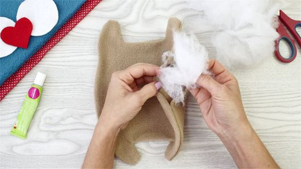 Hands filling a partially sewn plush toy with white polyester fiber stuffing on a crafting table with glue and scissors nearby.