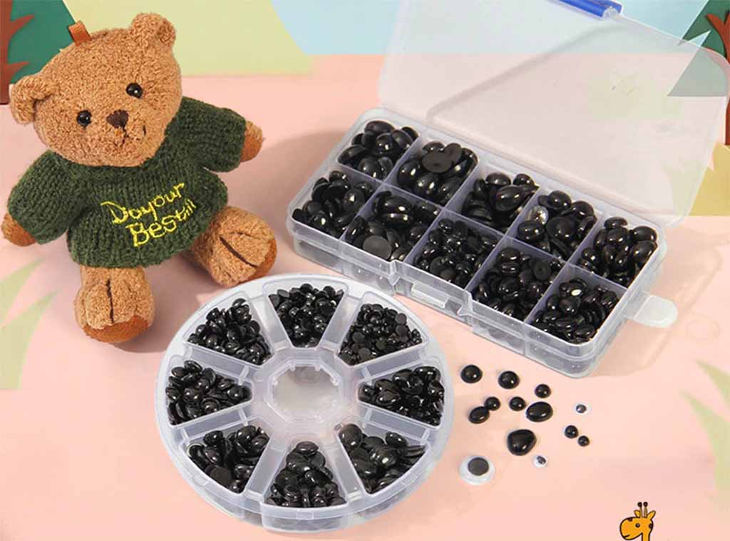 Assorted black plastic safety eyes and noses for plush toys displayed in organizer boxes beside a small teddy bear wearing a green sweater.