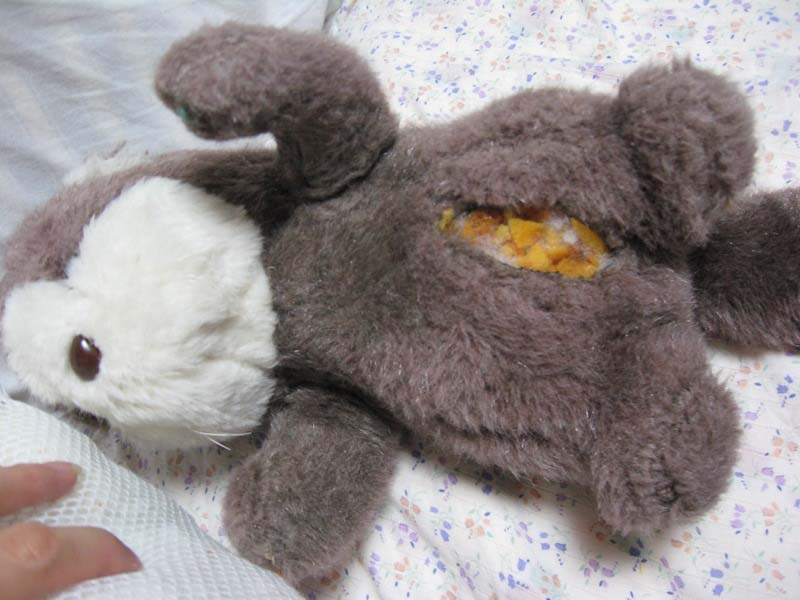 A brown plush animal with a large tear in its body, exposing the yellow and white stuffing inside, lying on a patterned bedsheet.