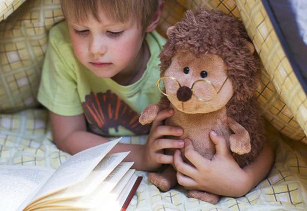 A young child lying under a blanket while reading a book and holding a soft brown hedgehog plush toy wearing tiny round glasses.