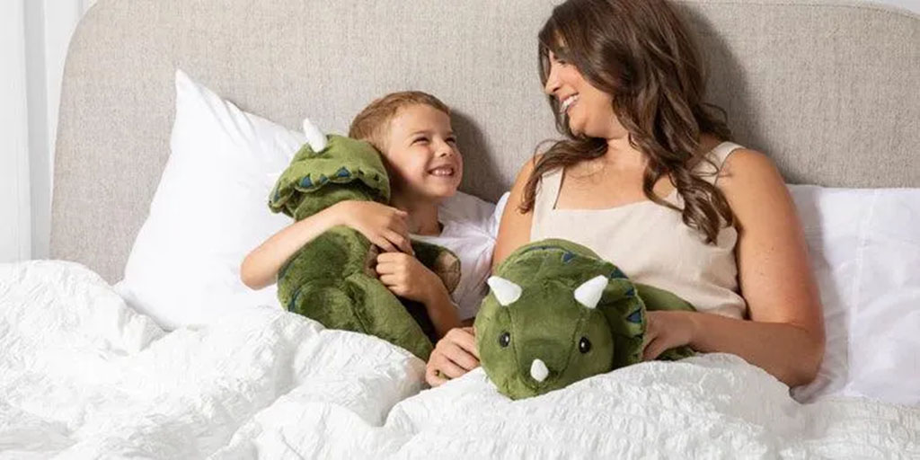 A mother and child smile at each other while sitting in bed, each holding a green dinosaur plush toy under a white blanket.
