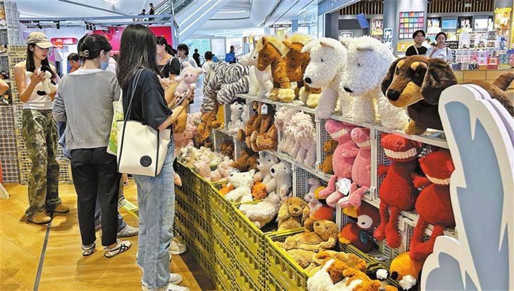 Shoppers browse a busy market stall filled with plush toys, including dogs, polar bears, rabbits, and colorful character plushies displayed on shelves and bins, with people examining and selecting items in a bright indoor mall setting.