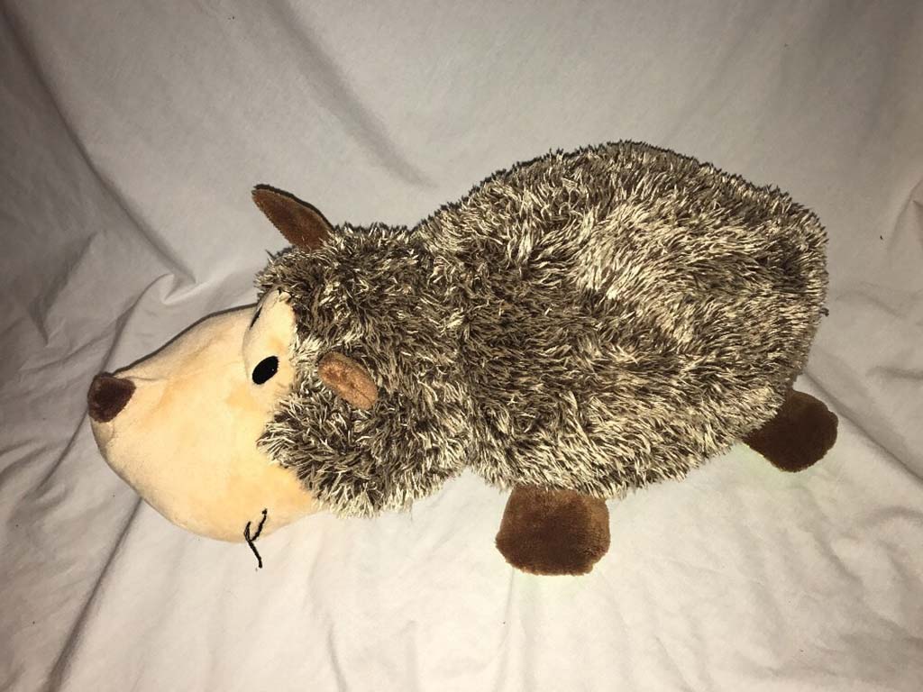 A cute hedgehog plush toy with a beige face, brown ears and paws, and a fuzzy gray body, displayed on a white fabric background.