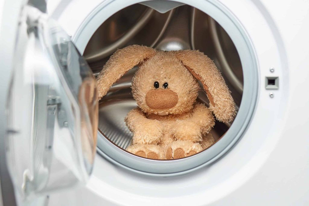 A soft brown bunny plush toy sitting inside a front-loading washing machine, ready to be cleaned.
